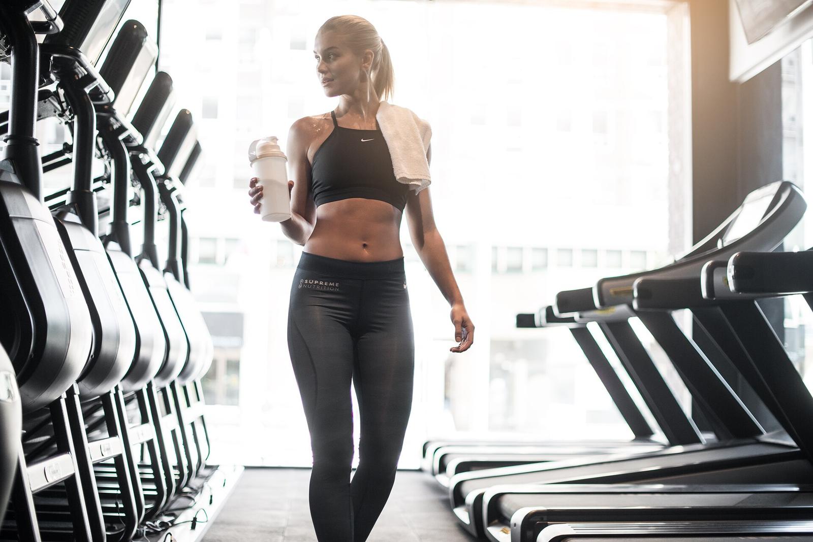 A woman holding a shaker bottle and towel as she walks past treadmills in a bright gym