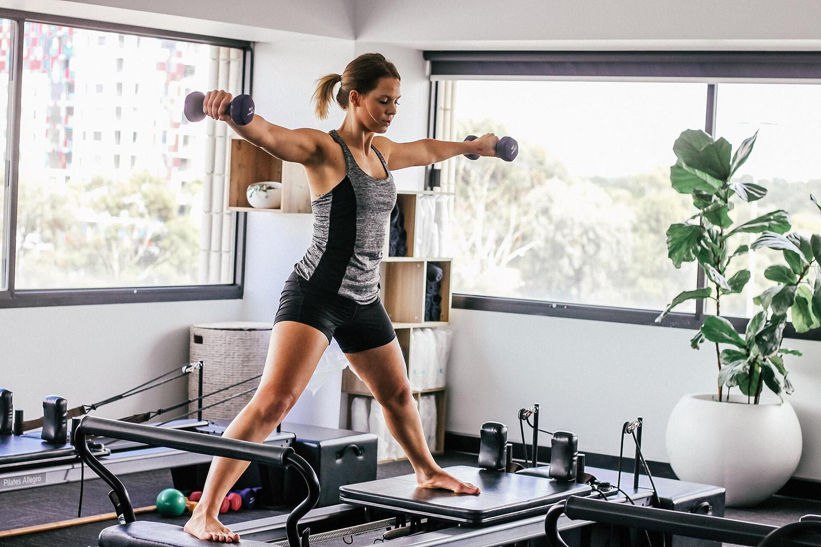 A woman lunging with dumbbells beside pilates reformers in a bright studio