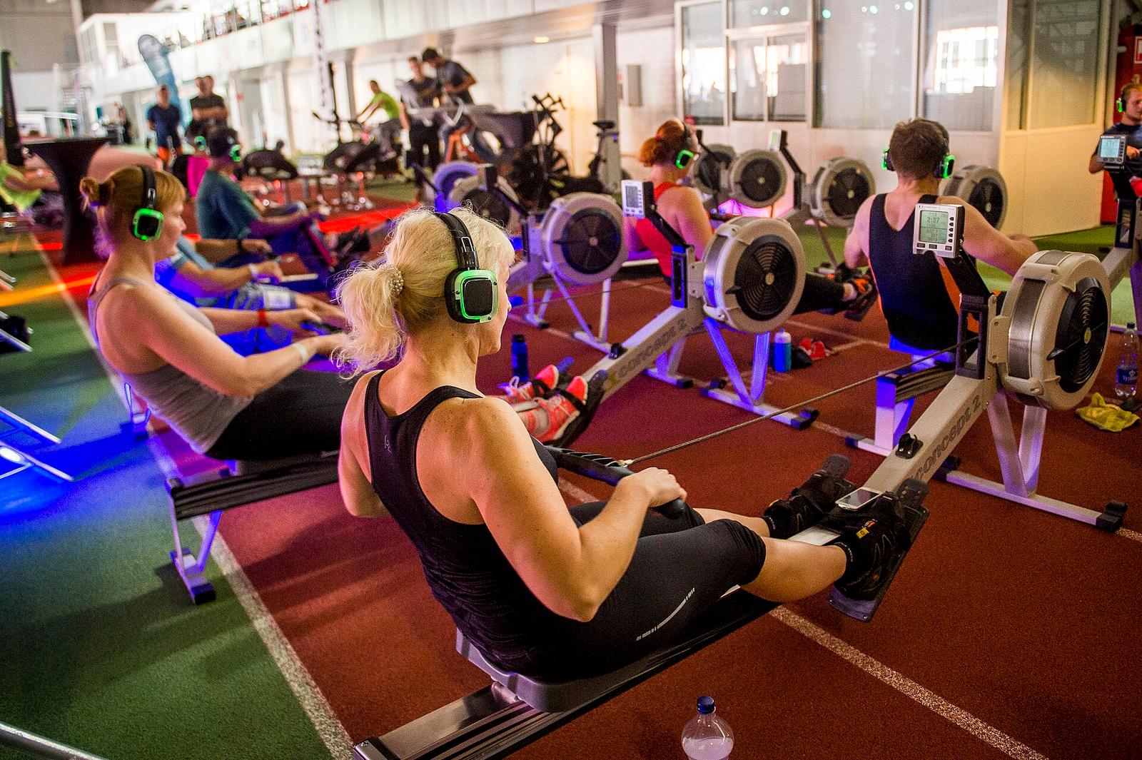 A group rowing class on indoor rowers, participants wearing wireless headphones under coloured lights